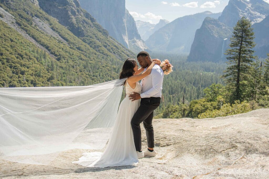 Couple embracing during a Yosemite elopement at Tunnel View, with the bride’s veil flowing in the wind and Yosemite Valley in the background.